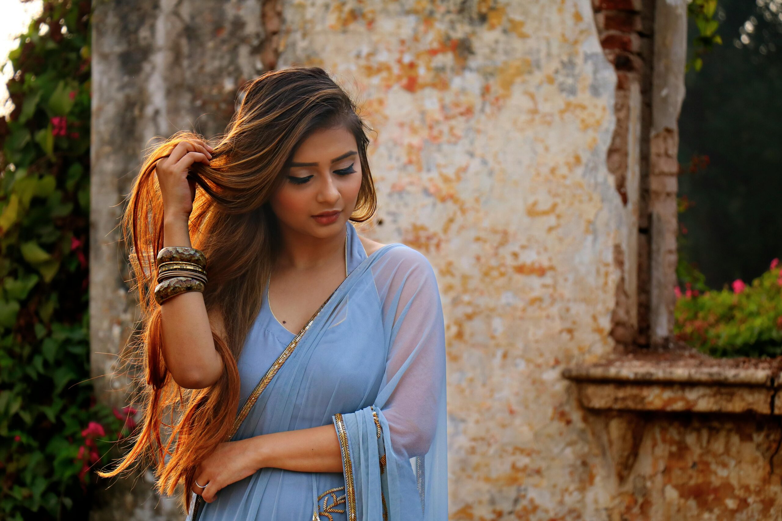 A woman in a blue saree poses gracefully outdoors by a rustic wall.